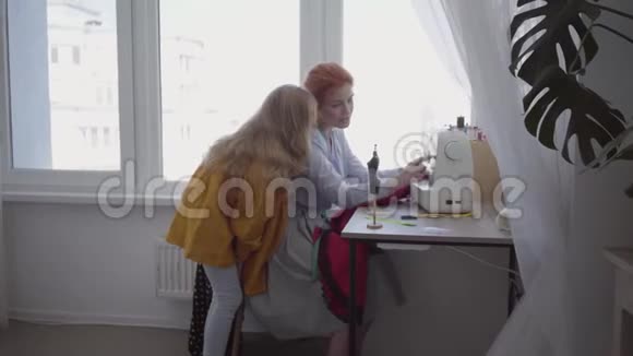 漂亮的红发妈妈把坐在靠窗的桌子旁的衣服缝好告诉她的女儿如何制作视频的预览图