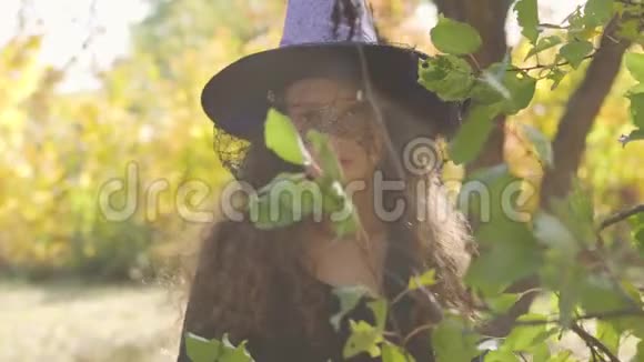 严肃的红发白种人女孩穿着万圣节女巫的服装站在绿叶后面的秋天公园里儿童视频的预览图