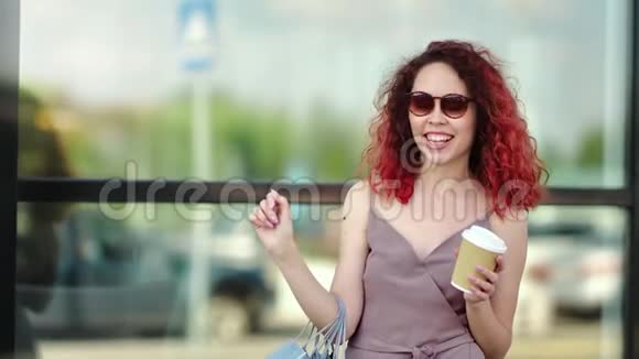 微笑的购物狂红发少女戴墨镜拿着咖啡纸杯视频的预览图