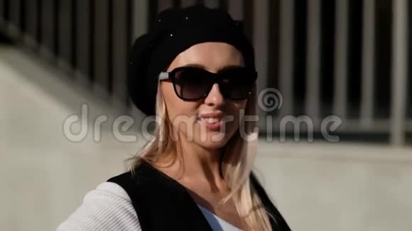 一位金发碧眼的美女戴着贝雷帽和和太阳镜看着相机微笑着在户外靠近视频的预览图