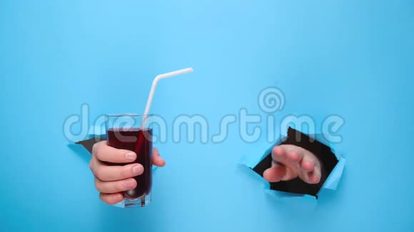 紧紧地握着一杯新鲜的红色果汁用吸管穿过撕裂的蓝色纸墙视频的预览图