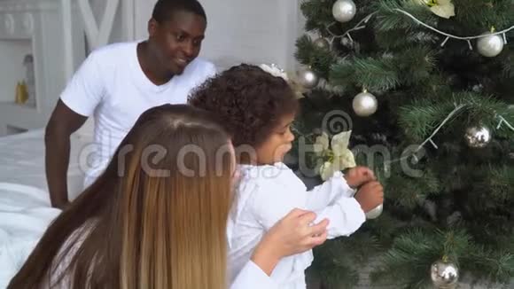 快乐的女婴在圣诞树上挂球孩子们装饰圣诞树年轻幸福的多民族家庭视频的预览图
