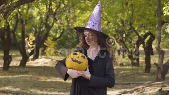 可爱的红发白种人女孩穿着万圣节女巫的服装站在秋天的公园里小孩抱着黄色南瓜视频的预览图