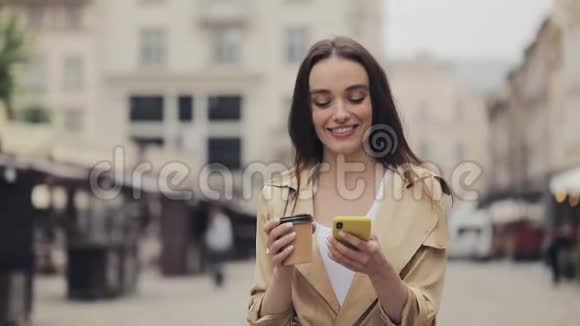 戴着鸽子沟的漂亮年轻女人用她的智能手机和微笑拿着纸杯喝着咖啡视频的预览图