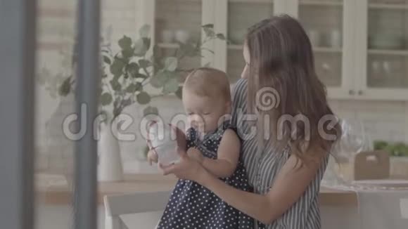 可爱的微笑的年轻女人用瓶子喂她的女婴厨房里有婴儿的假人幸福的概念视频的预览图