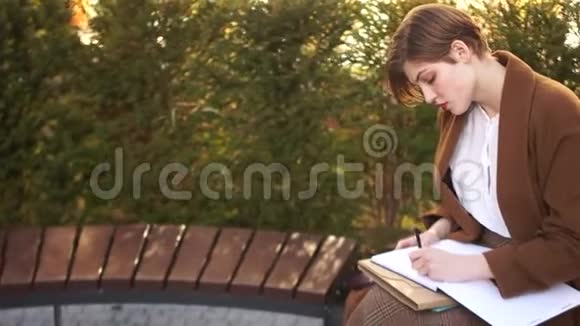 快乐学生轻松户外阅读和笔记簿的肖像短发女孩坐在公园的长凳上视频的预览图