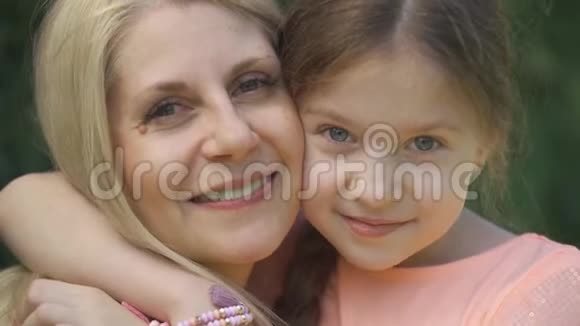 特写肖像微笑的金发母亲和她的小女儿拥抱在夏季公园看着相机快乐视频的预览图