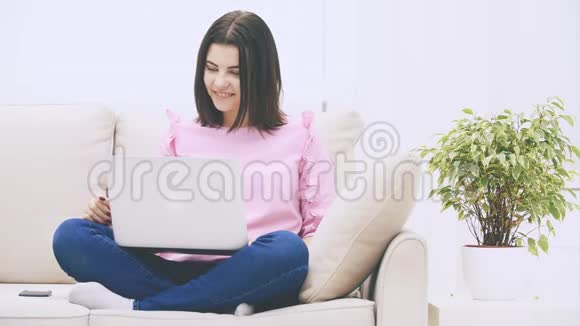 可爱的白种人黑发女孩坐在沙发上拿着笔记本电脑在互联网上冲浪气视频的预览图