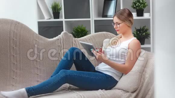 休闲专注的少女戴着时髦眼镜热衷于在家里坐在沙发上使用平板电脑视频的预览图