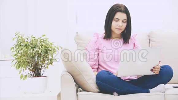 可爱的白种人黑发女孩坐在沙发上拿着笔记本电脑上网冲浪举止优雅视频的预览图