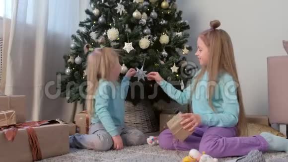 女孩们姐妹用礼物摇着盒子坐在圣诞树附近玩视频的预览图