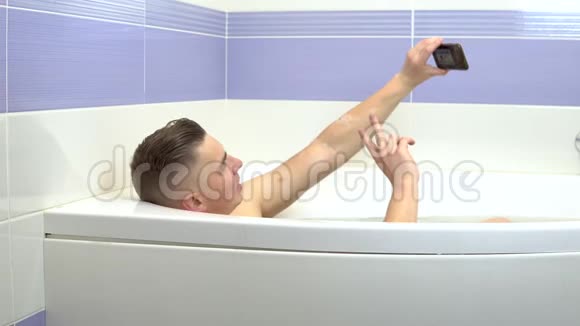 一个年轻人躺在浴缸里在电话里自拍男人在洗澡时放松视频的预览图