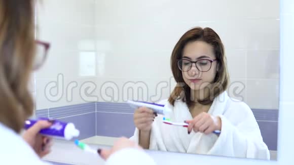 一个戴眼镜的年轻女人刷牙一个穿着白色外套的女人开始刷牙从镜子里看到视频的预览图