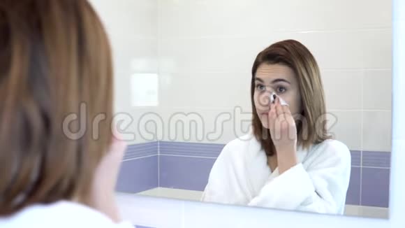 一个年轻的女人用棉垫从脸上卸妆一个穿着白色浴袍的女孩在浴室镜子前视频的预览图