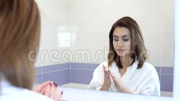一个年轻的女人在镜子前把面霜涂在脸上水疗程序浴室里穿着白色浴袍的女孩视频的预览图
