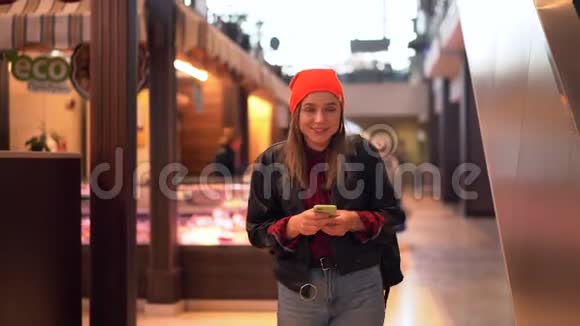 一个年轻漂亮的白人女孩穿着黑色皮夹克和牛仔裤走过购物中心看着视频的预览图