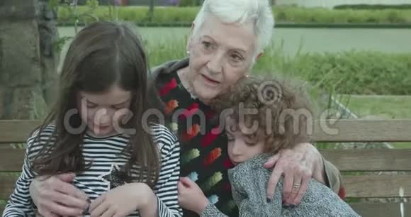 当两个小孩坐在公园的长凳上拥抱时一位年长的妇女拥抱并挤压着他们这个小男孩嬉戏地戳着女孩视频的预览图