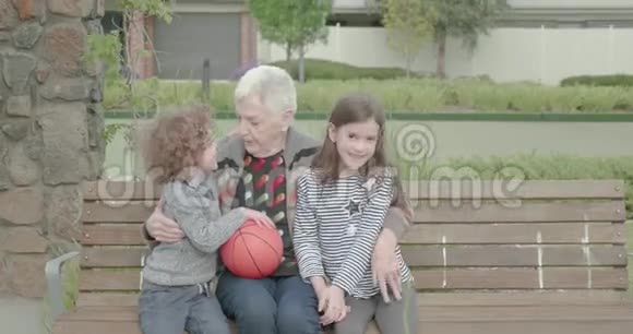 当两个小孩坐在公园的长凳上拥抱时一位年长的妇女拥抱并挤压着他们这位年轻的男孩正拿着一个篮球视频的预览图