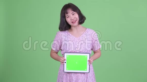 快乐的亚洲孕妇一边说话一边显示数字平板电脑视频的预览图