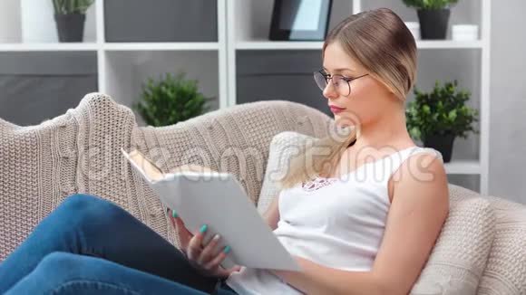 时尚眼镜中的漂亮女孩专注于阅读有趣的纸质书在沙发上放松视频的预览图