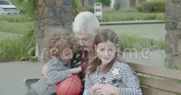 当两个小孩坐在公园的长凳上时一位老妇人抱着一个篮球挠痒痒视频的预览图