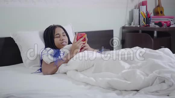 镜头接近非洲裔美国女孩与可怕的躺在白色床上和使用智能手机年轻女子冲浪视频的预览图