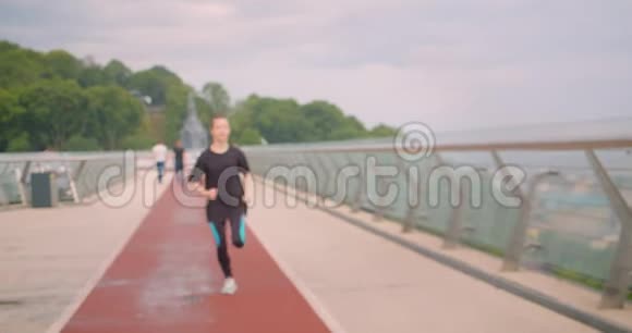 穿着黑色t恤在城市户外的桥上奔跑的年轻白种人运动型女性慢跑者的特写照片视频的预览图