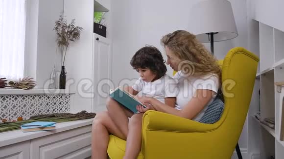 迷人的微笑男孩和他的金发无忧无虑的迷人母亲坐在舒适的椅子上一起看书视频的预览图