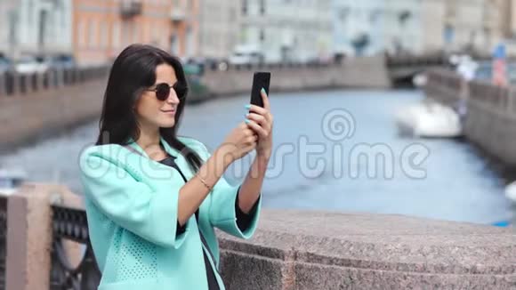 可爱的时尚女孩用智能手机从历史城市景观堤拍照视频的预览图