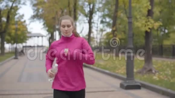 一位穿着粉红色运动服的白种人年轻女子走近镜头喝水和向前奔跑的肖像视频的预览图