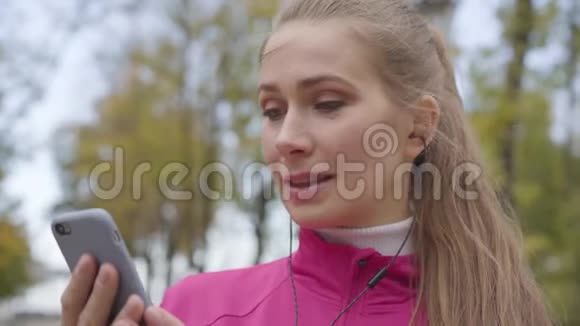 身着粉色运动服的白种人年轻女子的近脸戴上耳机打开音乐视频的预览图