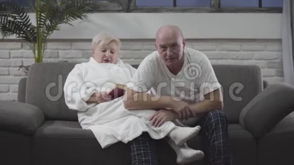 成熟的白人男人和妻子坐在沙发上看电视上的体育比赛女人在看书老年人休息视频的预览图