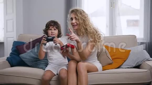 漂亮快乐快乐的妈妈和活泼可爱的小男孩坐在柔软的沙发上玩着游戏杆视频的预览图