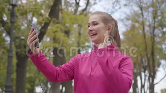 身着粉色运动服面带微笑的白人女运动员通过视频聊天聊天和竖起大拇指的照片开朗的视频的预览图