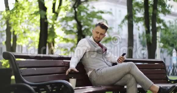 一个穿着昂贵西装的商人坐在公园长凳上在手机上读信息视频的预览图