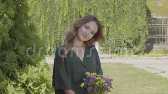 一幅俏皮可爱的年轻女子拿着一束野花站在户外看着相机的肖像概念视频的预览图