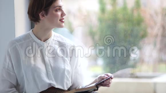 可爱的年轻女人坐在她家的窗户上做梦她手里拿着一本打开的书视频的预览图