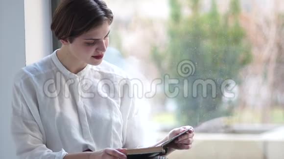 一个穿着一件老式白色衬衫的年轻女孩的亲密Indore肖像一个女人坐在窗前看书视频的预览图