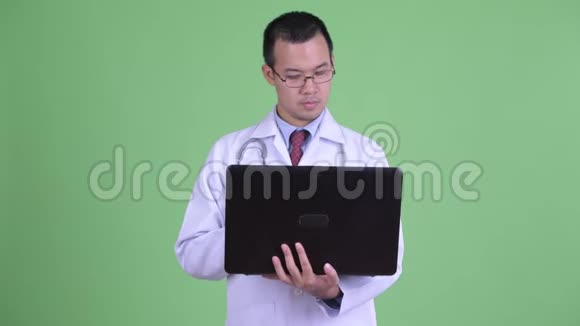 快乐的亚洲男子医生一边思考一边说话一边使用笔记本电脑视频的预览图