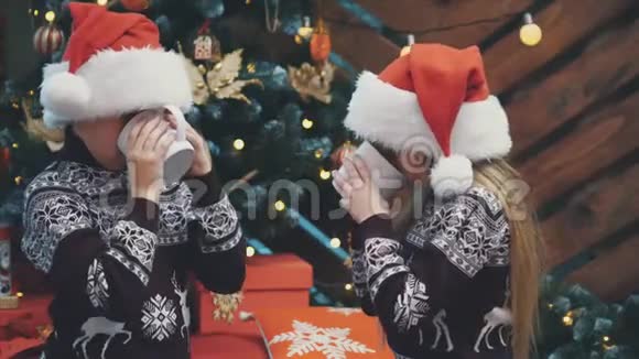 戴着圣诞帽和羊毛衫的漂亮孩子坐在圣诞树旁手里拿着一杯可可看上去很高兴视频的预览图
