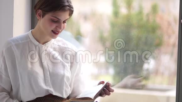 留着短发的年轻女孩坐在窗边手里拿着一本书看着相机微笑着视频的预览图