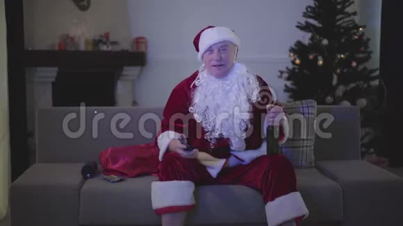 穿着圣诞老人服装坐在沙发上喝啤酒在家看电视的积极白人的肖像高级官员视频的预览图