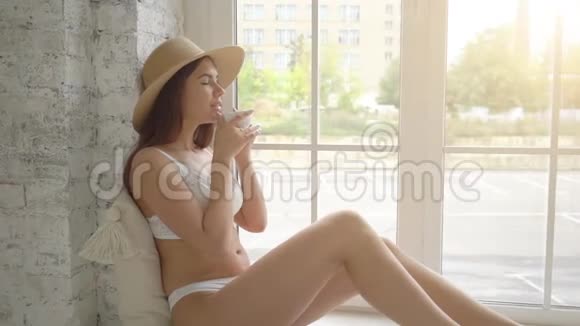 漂亮的女青年戴着草帽在家坐在窗台上喝早咖啡视频的预览图