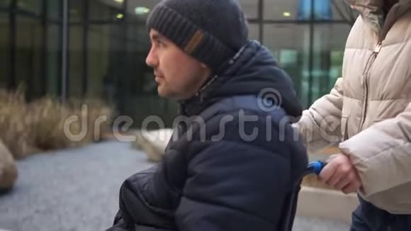 穿着白色夹克的女人拿着一个残疾的年轻人坐在轮椅上那人挥动他的手支助和援助视频的预览图