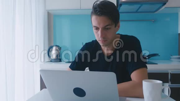 年轻的惊讶男子震惊和惊讶他看到在笔记本电脑时坐在家里的厨房桌子视频的预览图