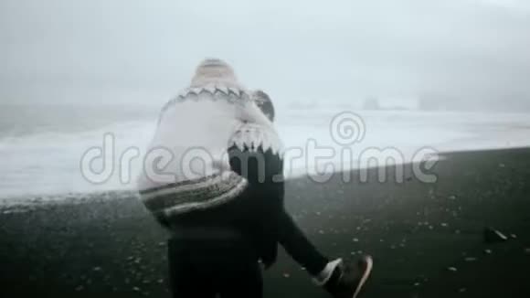年轻的幸福夫妇在黑色海滩的岸边玩得很开心女人跳到男人背上拥抱他视频的预览图