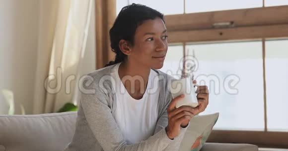冷静微笑的非洲女人坐在沙发上喝着咖啡做梦视频的预览图