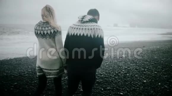在冰岛黑色海滩上散步的年轻漂亮夫妇的后景手牵着手说话视频的预览图