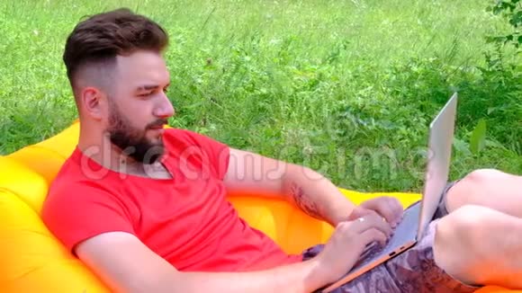 一个穿红色T恤的年轻人躺在空气沙发上在笔记本电脑上工作有胡子的人做生意视频的预览图