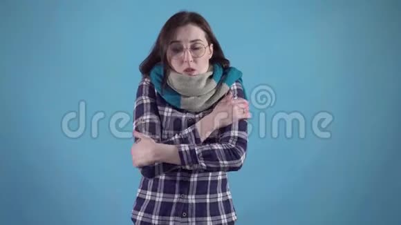 戴着围巾和眼镜的冻僵的年轻女人站在蓝色的背景上不会感到温暖视频的预览图